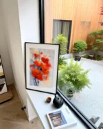 Tablou cu flori rosii aprinse pe un pervaz, langa o fereastra mare cu vedere la o gradina cu plante verzi